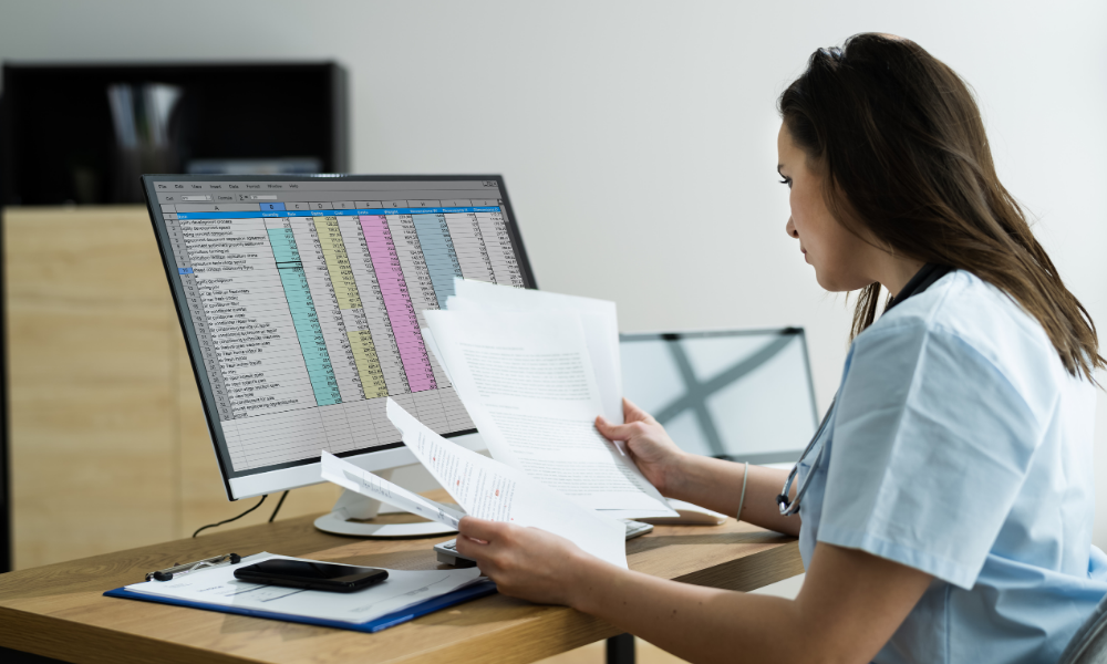 Female professional reviewing printed documents while working with a large event registration spreadsheet on a desktop computer—illustrating the challenges of manual attendee tracking before using Salesforce-native event management.