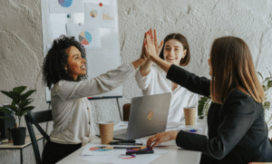 Three event marketing professionals high-five in a modern office while collaborating on event branding strategy using data charts and Salesforce-based tools
